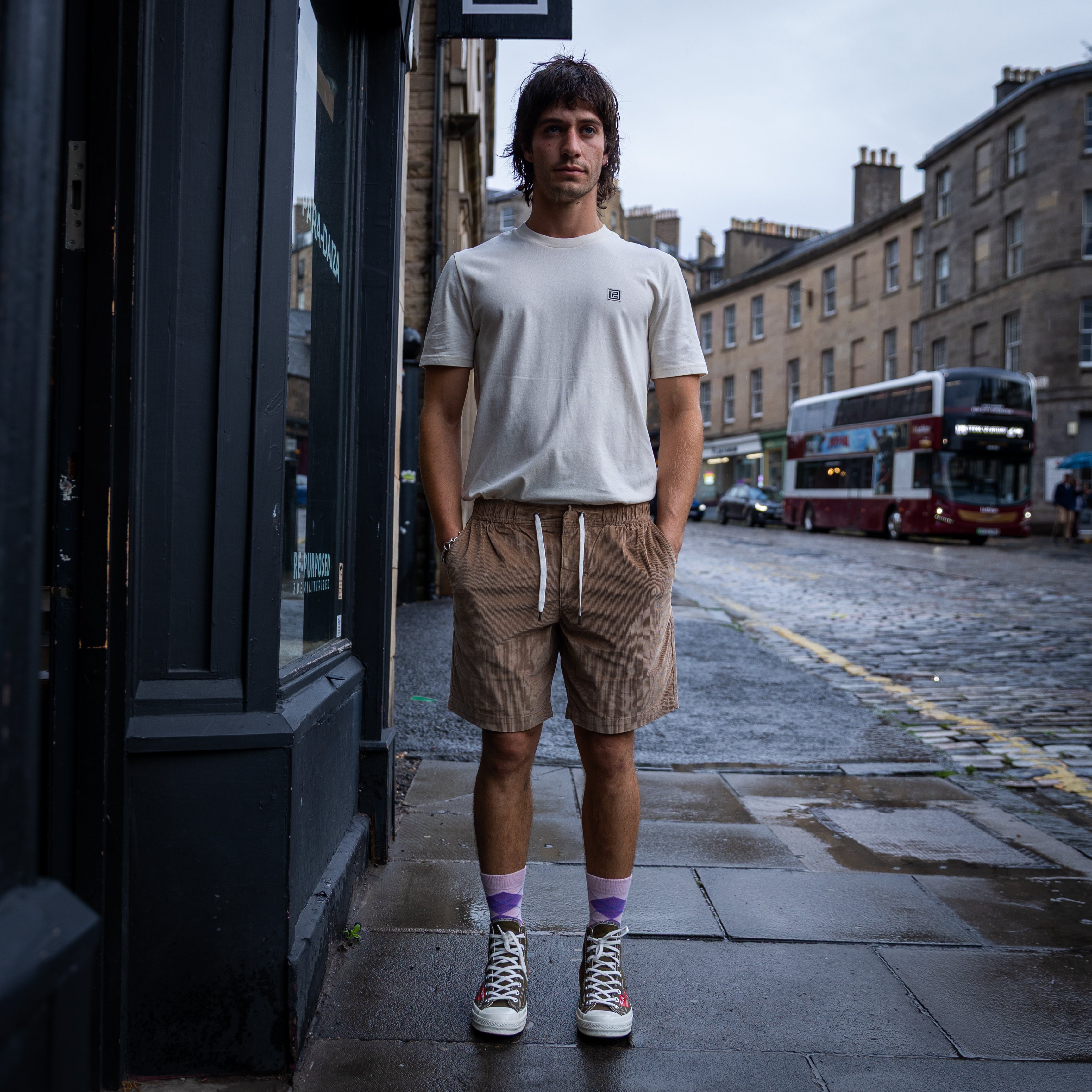 A man stands on a city sidewalk wearing a white crew-neck t-shirt with a small chest logo, tan drawstring shorts, purple socks, and white high-top sneakers. The background shows a wet street with brick buildings and a double-decker bus.