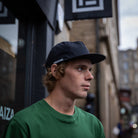 A man wearing a dark navy blue Paradaiza baseball cap and a green t-shirt, standing outdoors on a city street with blurred buildings in the background.