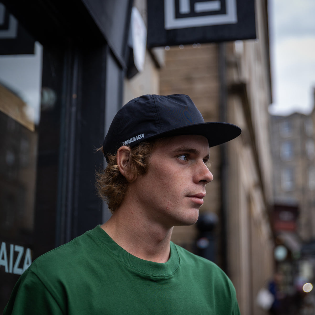 A man wearing a dark navy blue Paradaiza baseball cap and a green t-shirt, standing outdoors on a city street with blurred buildings in the background.