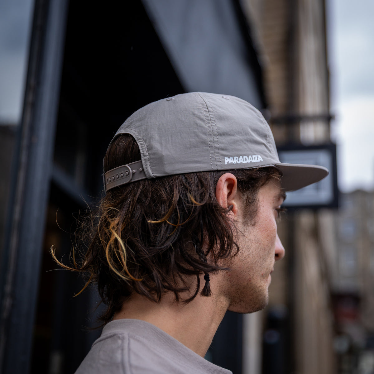 A man in profile wearing a light gray Paracasa baseball cap with a structured crown and adjustable strap, set against a blurred urban background.