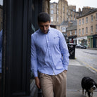 A man wearing a light blue long-sleeve button-down shirt and beige trousers walks on a city street, with a black and white dog in the background.