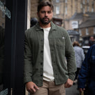 A man wearing an olive green button-up overshirt over a white t-shirt and beige trousers, standing on a city street.