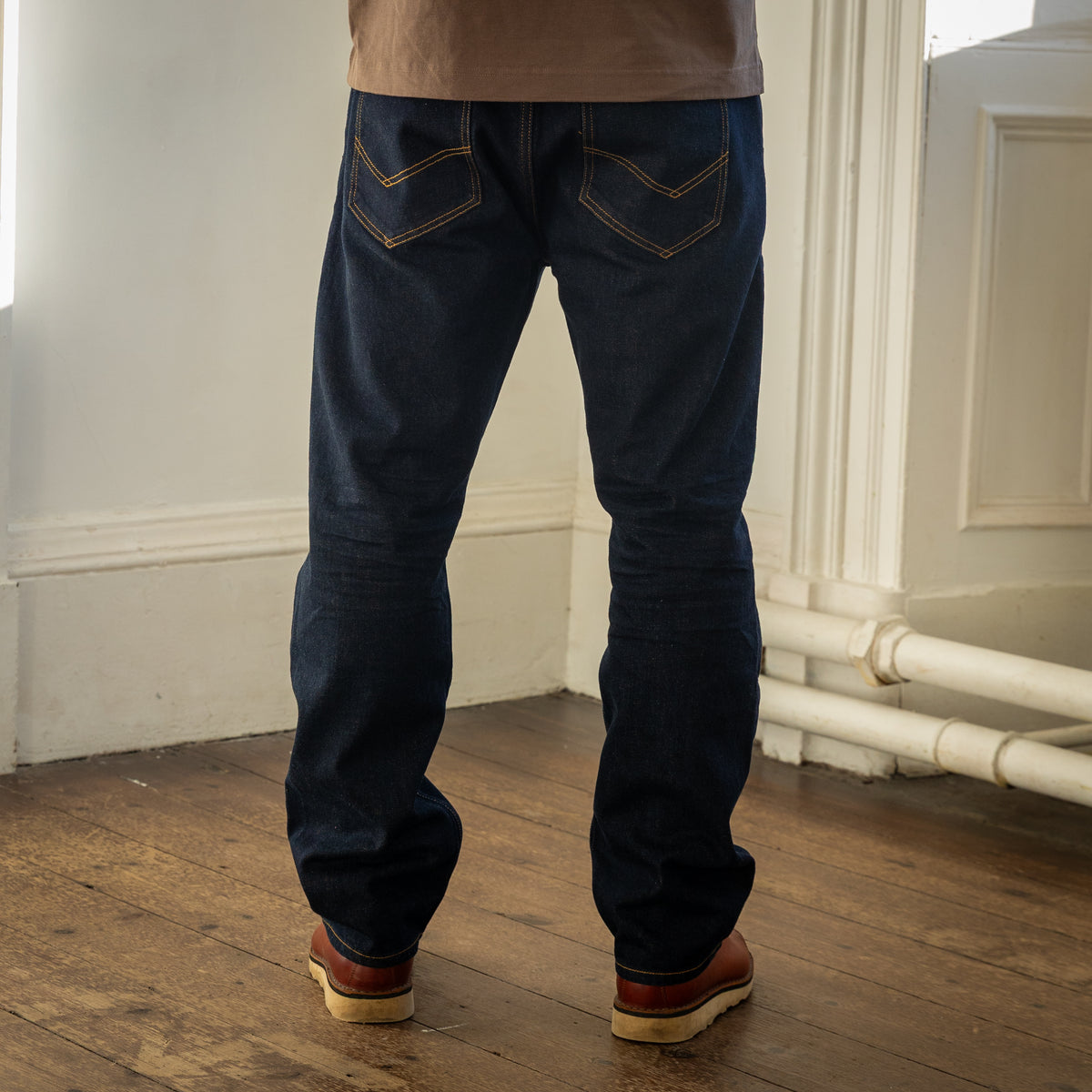 Rear view of a person wearing dark blue denim jeans with contrast stitching, paired with a tan top and brown shoes, standing on a wooden floor in an indoor room.