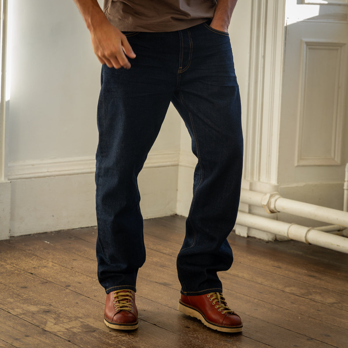 A person wearing dark indigo blue jeans and brown leather lace-up shoes, standing on a wooden floor in an indoor room with white walls and molding.