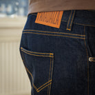 Close-up of the back waistband and pocket of dark indigo denim jeans with yellow contrast stitching, showing a tan leather patch with the brand name 'TANAJAZA' and a brown shirt hem.