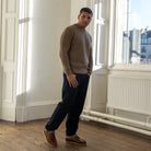 A man wearing a ribbed taupe mock-neck sweater, dark navy trousers, and brown leather shoes, standing in a bright room with white walls, wooden floors, and large windows.