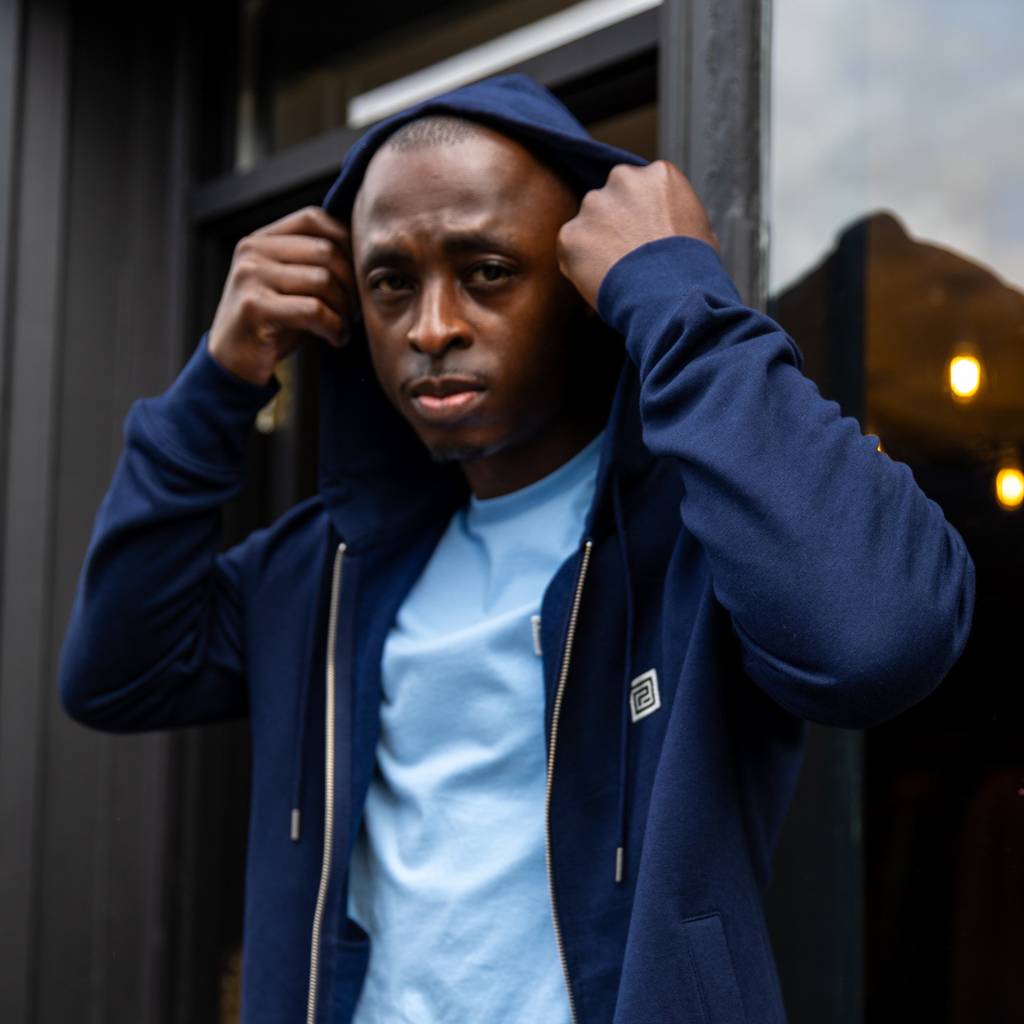 Man wearing a navy paradaiza hoodie with a brand logo, standing in front of a store window.