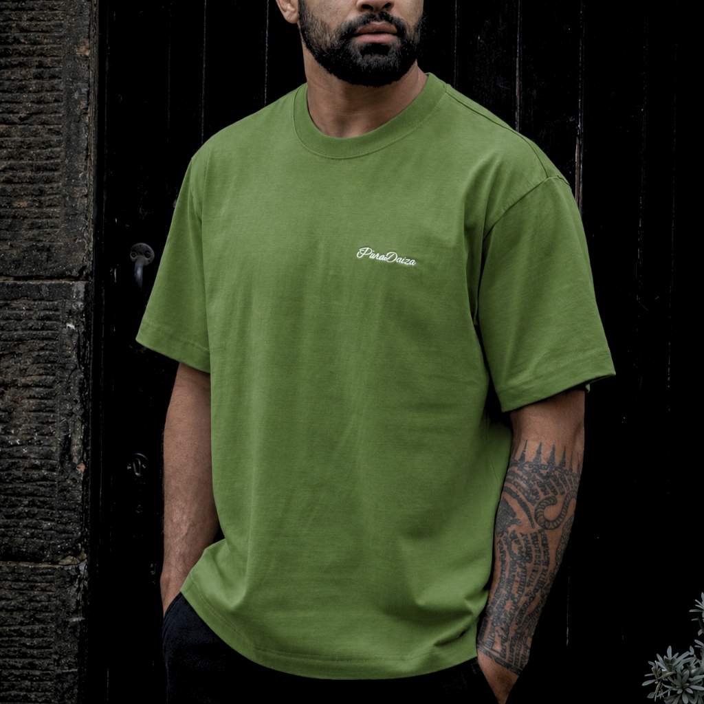 A man wearing green crewneck t-shirt with a small white embroidered 'PineDesire' logo on the left chest, standing against a dark wooden door.