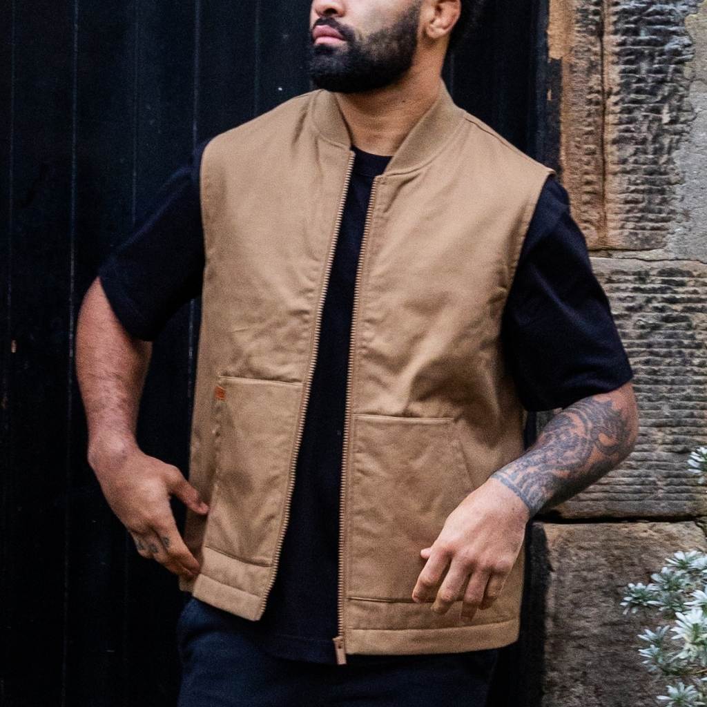 Man wearing a paradaiza tan vest over a black shirt standing in front of a dark door.
