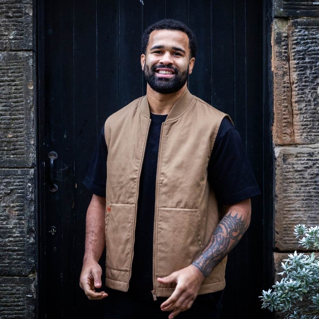 Man wearing a paradaiza tan vest over a black shirt standing in front of a dark door.