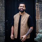 Man wearing a paradaiza tan vest over a black shirt standing in front of a dark door.
