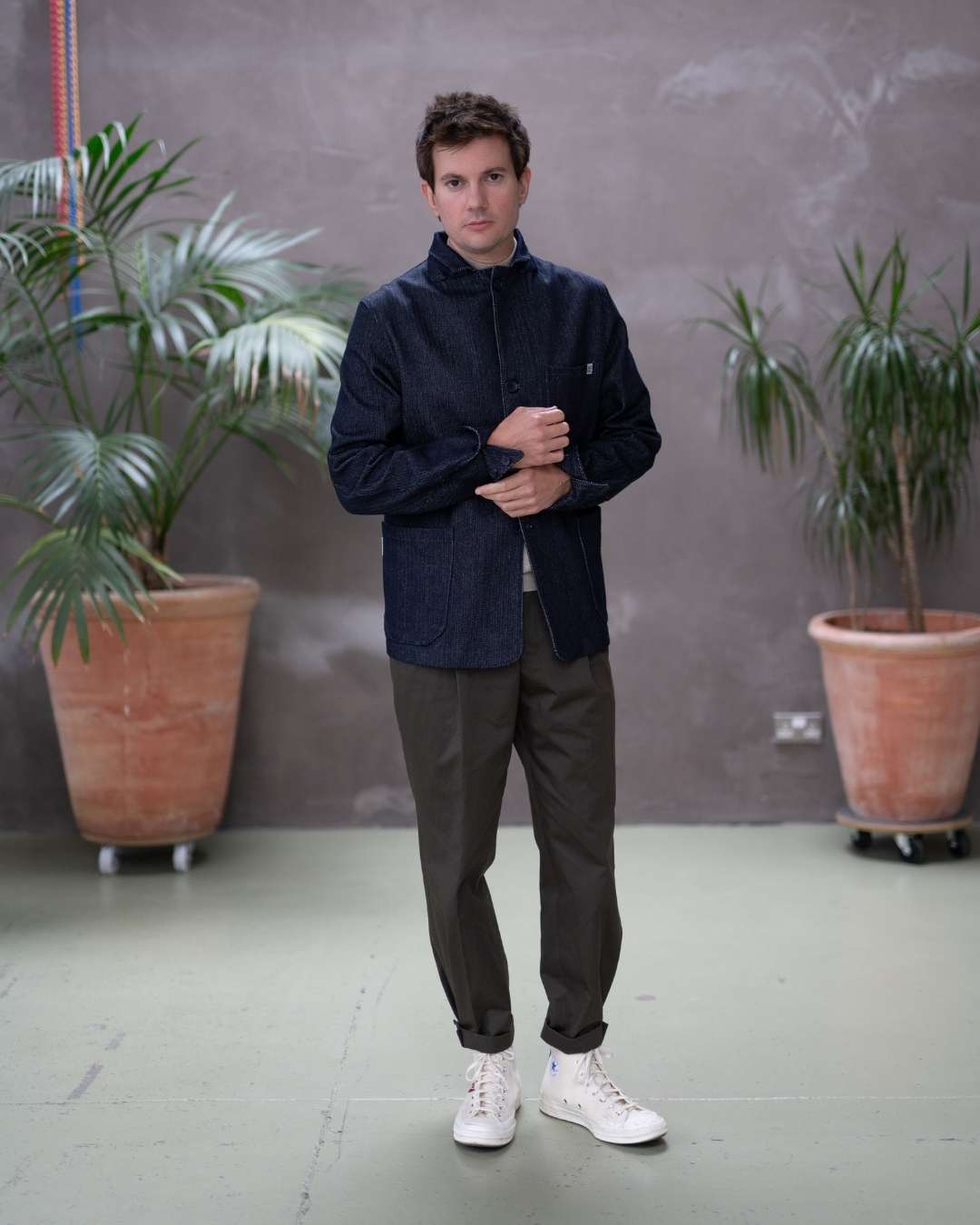 Model  wearing a dark denim paradaiza  jacket and khaki pinch chinos  standing indoors with plants in the background.