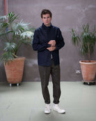 Model  wearing a dark denim paradaiza  jacket and khaki pinch chinos  standing indoors with plants in the background.