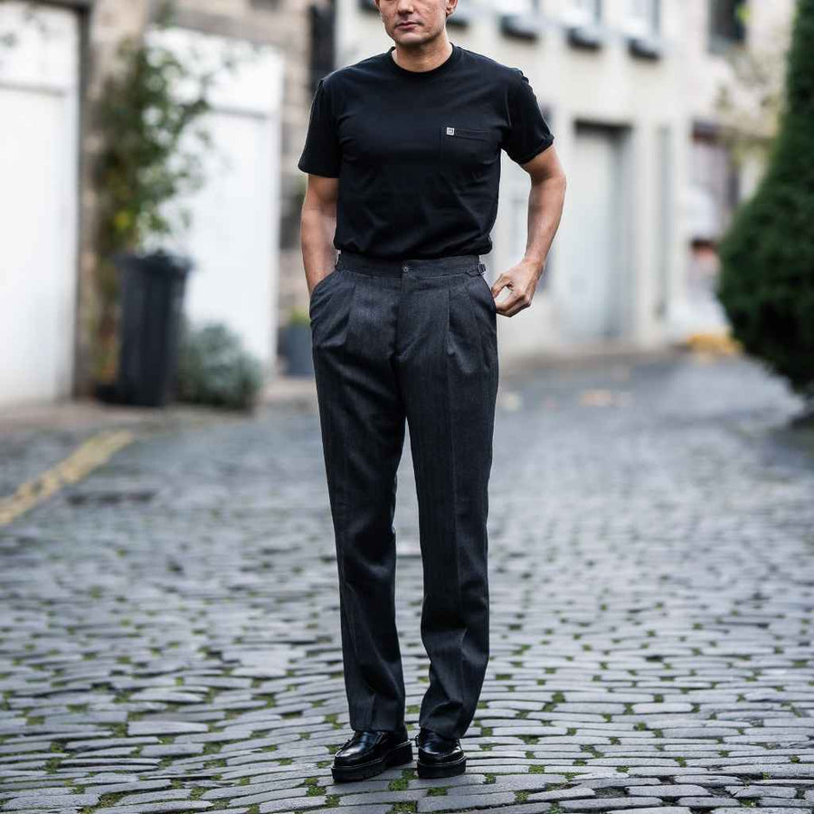 A man standing on a cobblestone street wearing paradaiza grey trousers and a black t-shirt.
