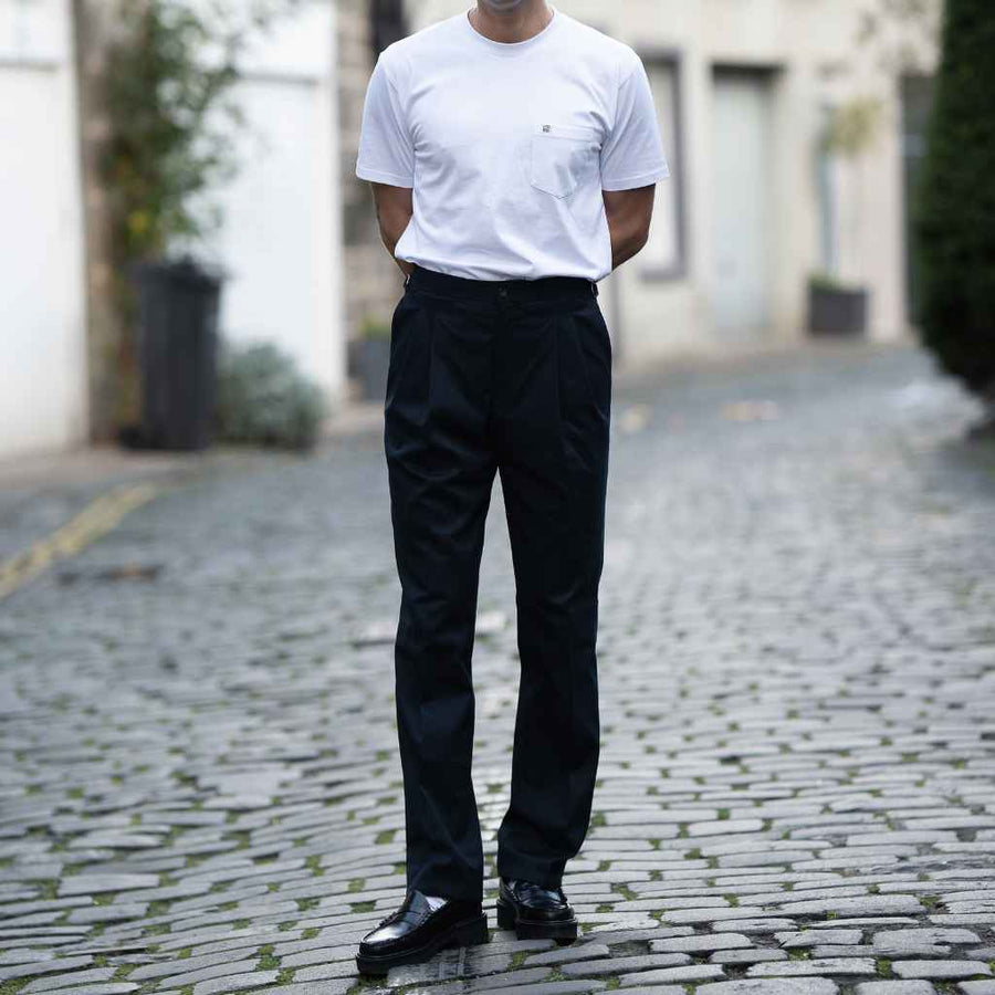 A person standing on a cobblestone street wearing paradaiza navy trousers and a white t-shirt.