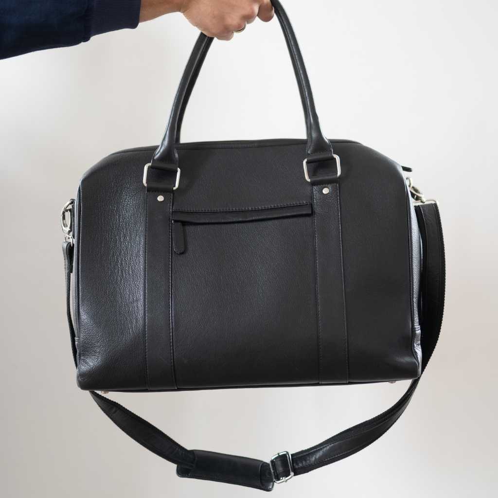 Man holding a  paradaiza black leather weekend bag in a room with a window .