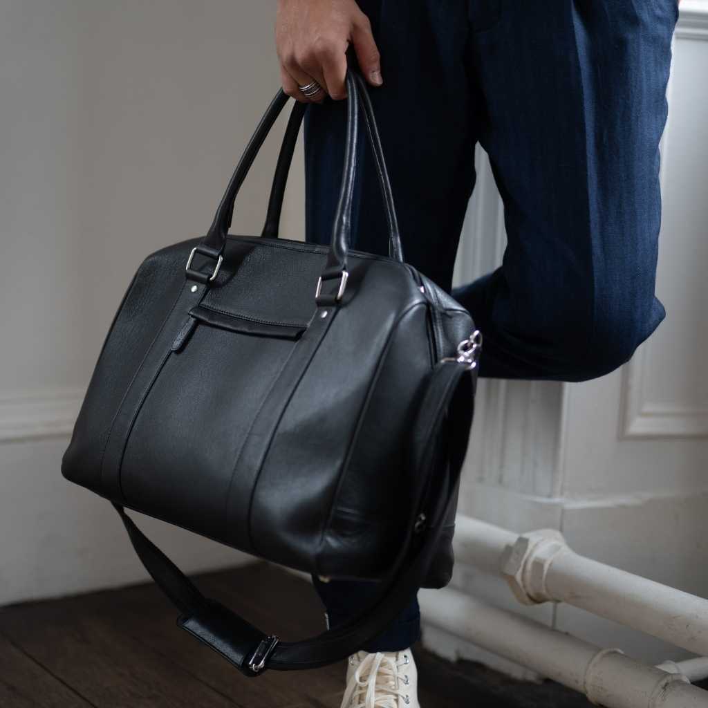 Man holding a  paradaiza black leather weekend bag in a room with a window .