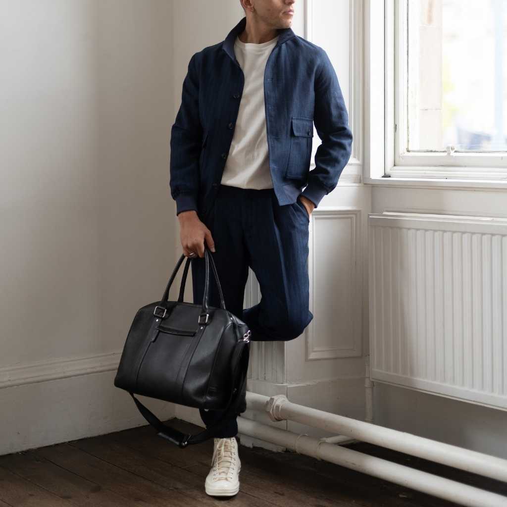 Man holding a  paradaiza black leather weekend bag in a room with a window .