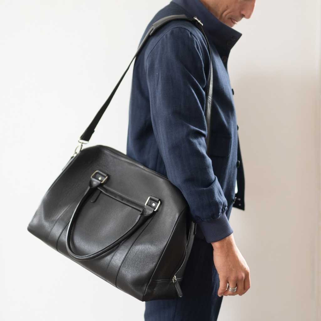 Man holding a  paradaiza black leather weekend bag in a room with a window .