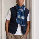 A man wearing a navy blue quilted vest over a white t-shirt and khaki pants, with a blue and white patterned scarf wrapped around his neck, standing indoors against a white wall.