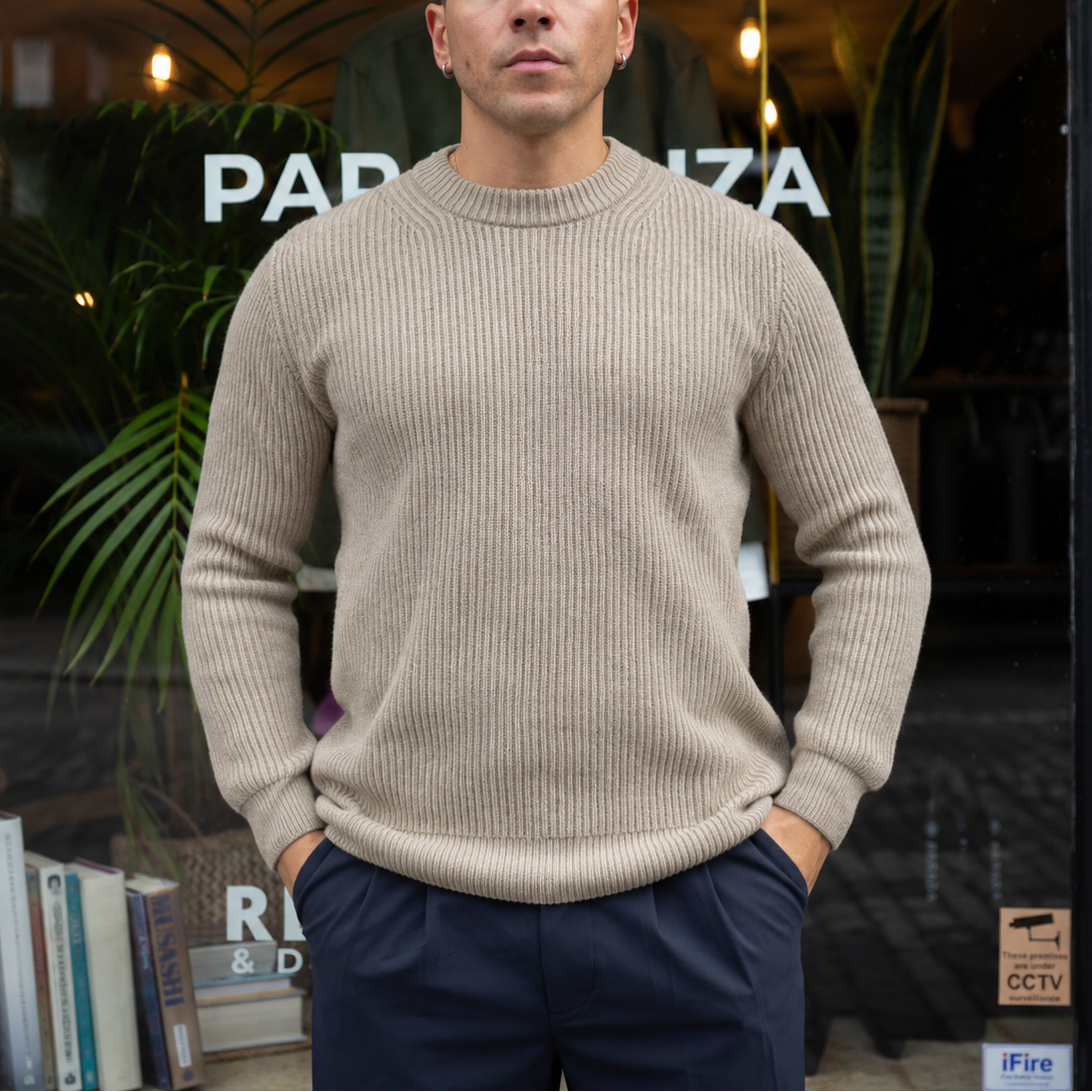 A man wearing a beige ribbed knit crewneck sweater and navy blue trousers, standing in front of a storefront window with plants and books visible in the background.