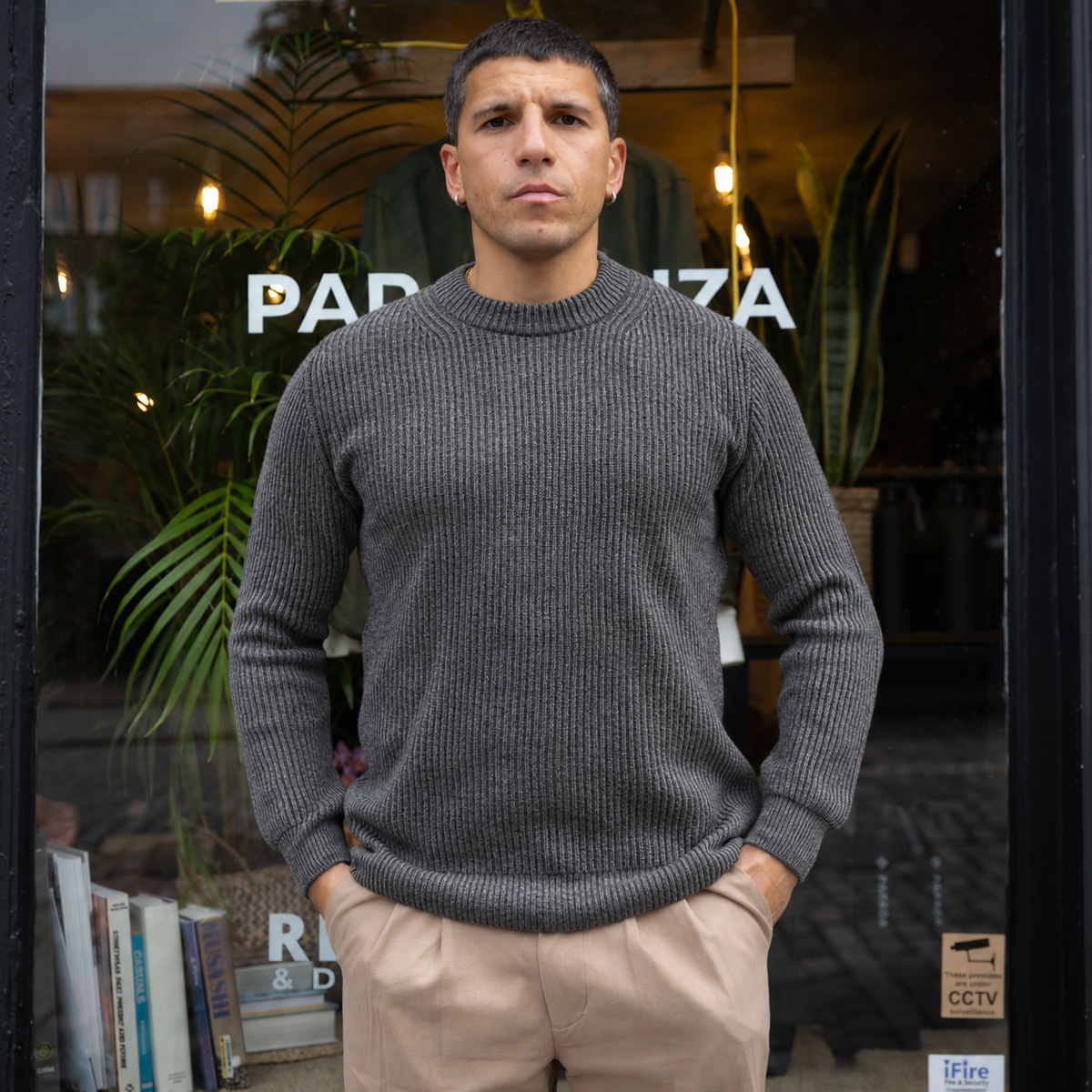 A man wearing a dark gray ribbed knit crewneck sweater and beige trousers, standing in front of a storefront window with plants and warm lights visible in the background.
