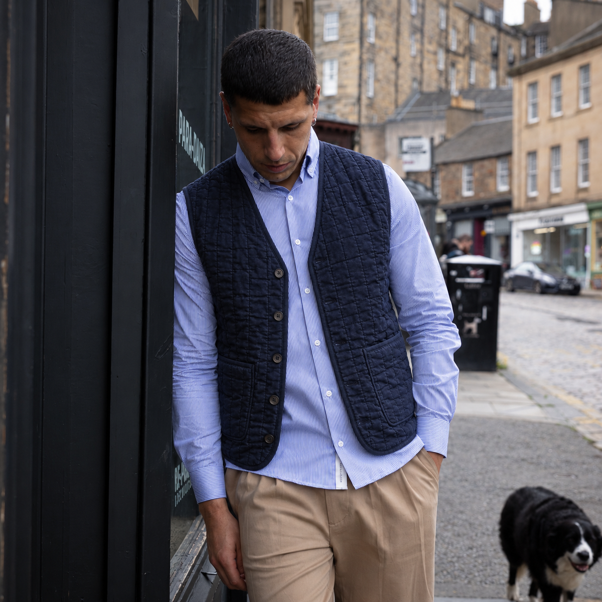 A man wearing a dark navy quilted button-up vest over a light blue dress shirt and khaki trousers, standing on a city street next to a black storefront with a dog in the background.