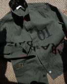 An Khaki  green utility jacket laid flat on a textured woven surface, featuring a large black '01' print on the chest, dark buttons, and red embroidered details on the pockets.