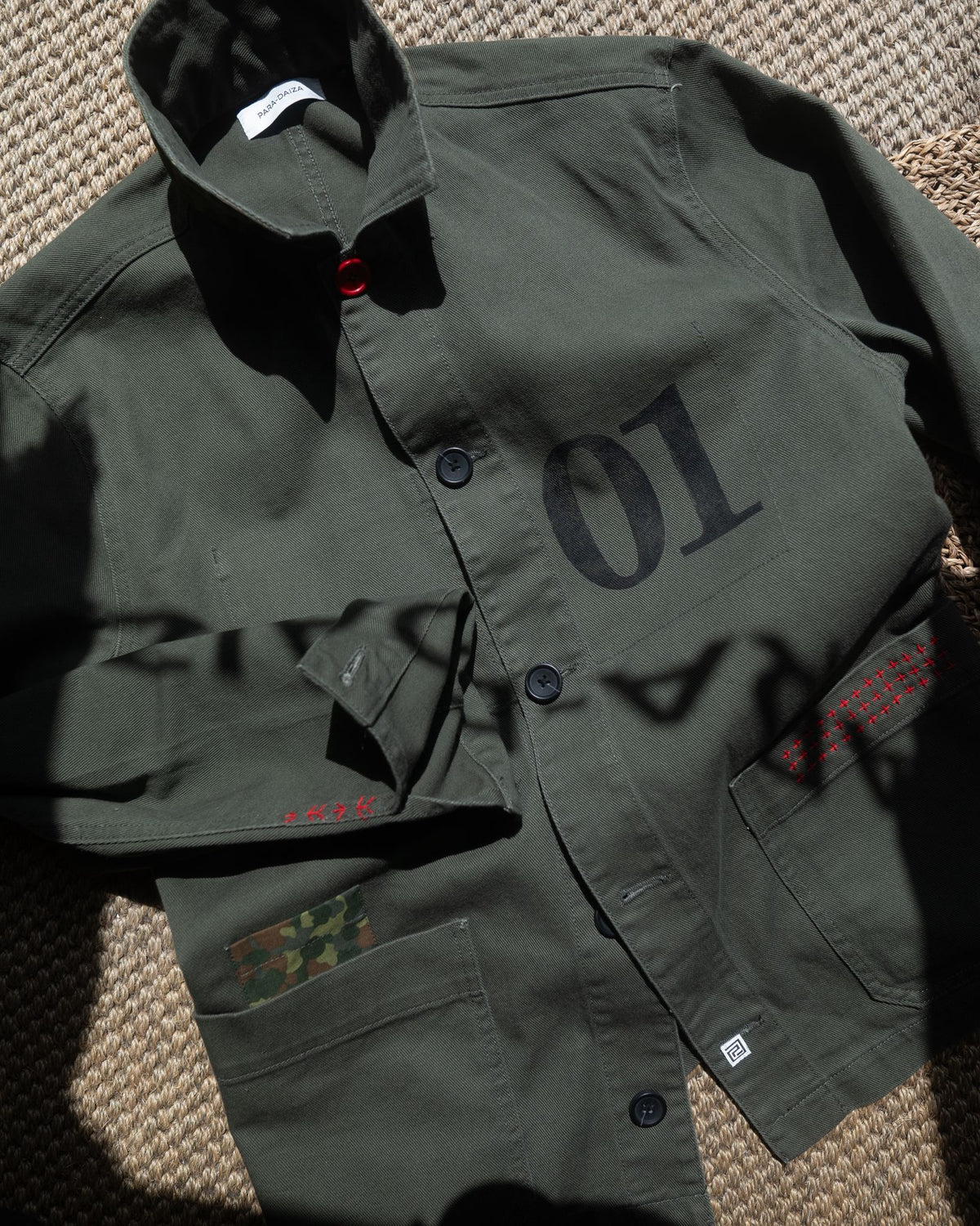 An Khaki  green utility jacket laid flat on a textured woven surface, featuring a large black '01' print on the chest, dark buttons, and red embroidered details on the pockets.