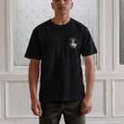 Man wearing a paradaiza  black t-shirt with a logo against a white paneled wall.