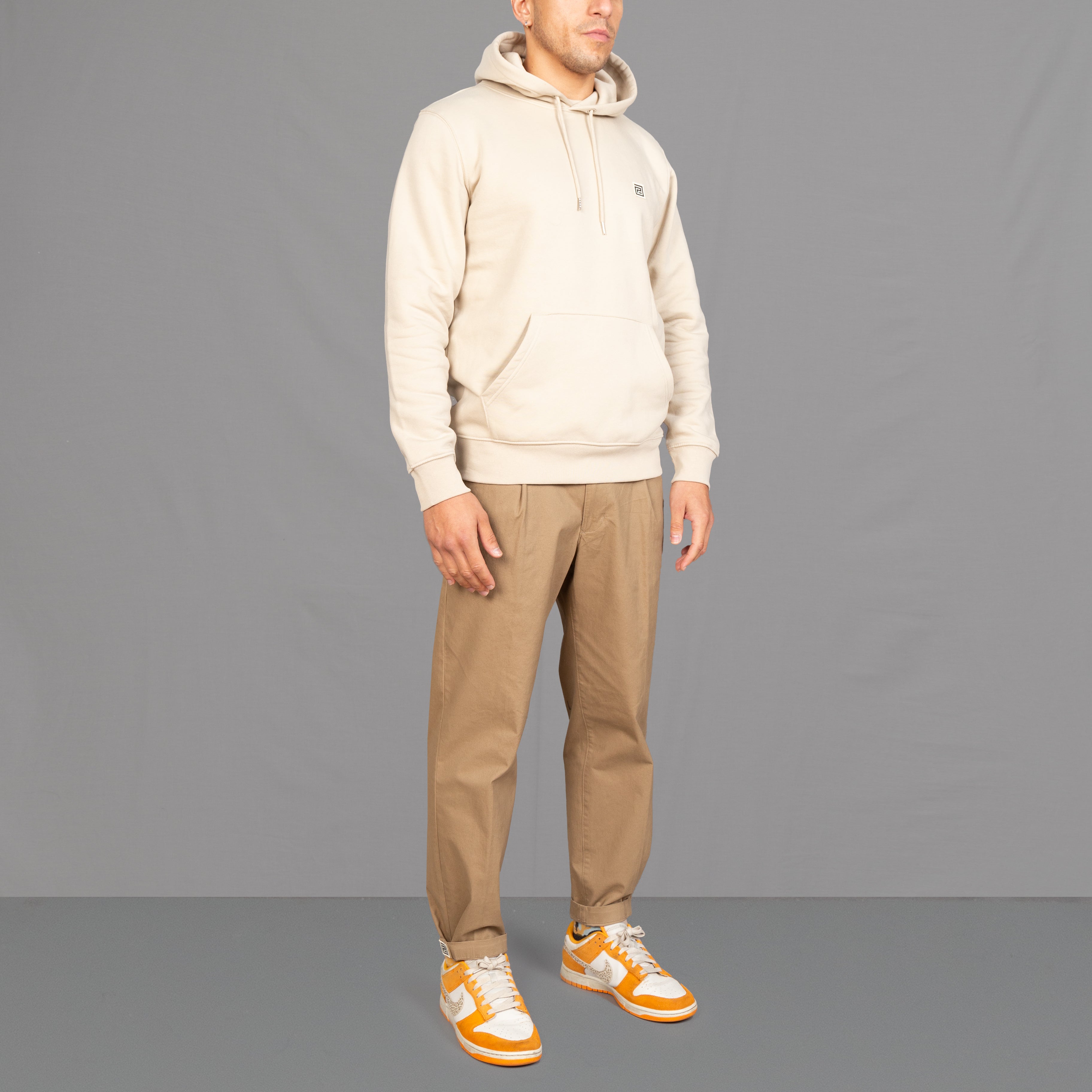 Model wears a sand colour hooded sweatshirt and tapered chino, with Nikes