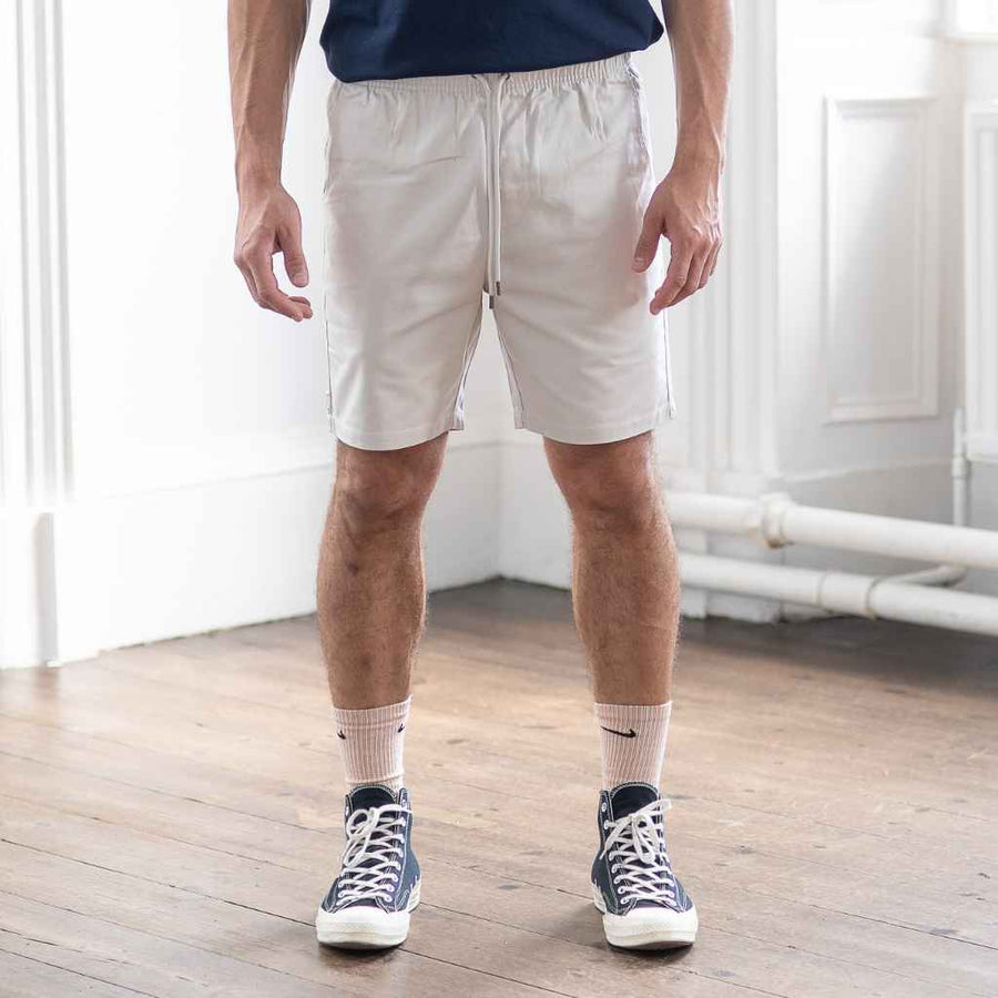 A person wearing paradazia white cotton shorts and a navy blue t-shirt standing in a room with wooden flooring.