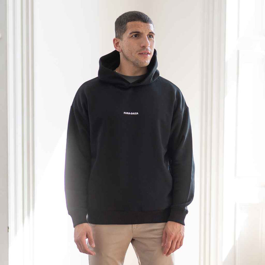A male model wearing a black Paradaiza oversized hooded sweatshirt with a visible brand logo on the chest area.