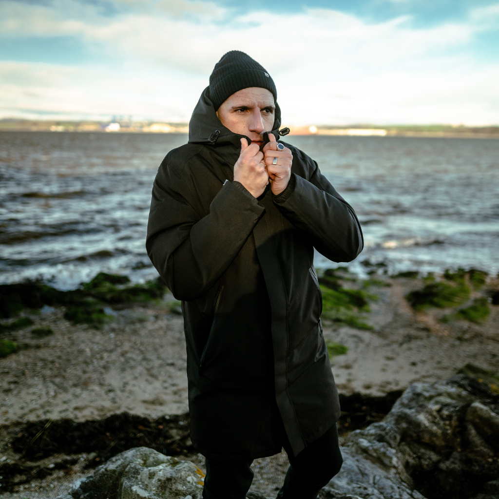 A man wearing a paradaiza black padded parka jacket with a hood, paired with a red sweater and a green cap, standing in a outdoor setting with grass and hills in the background.