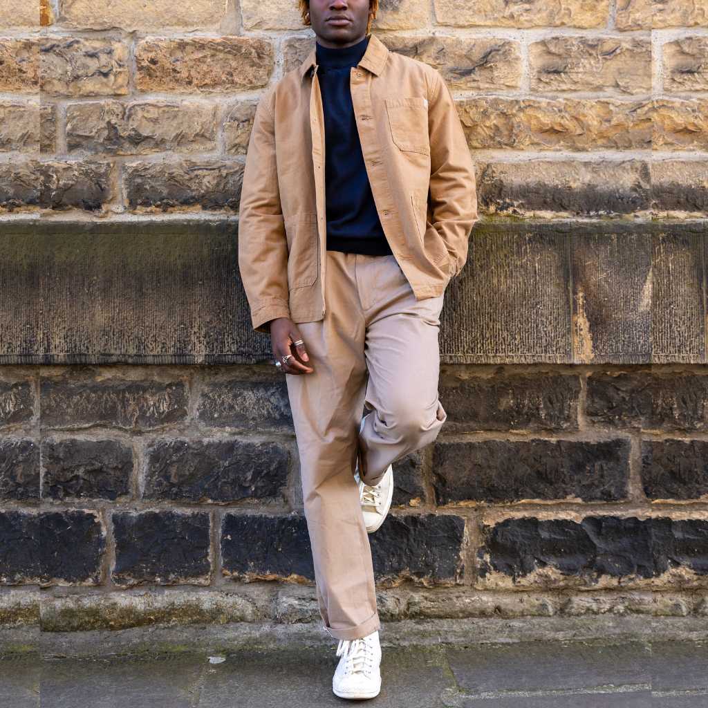 Person wearing a tan paradaiza jacket and pants leaning against a stone wall.