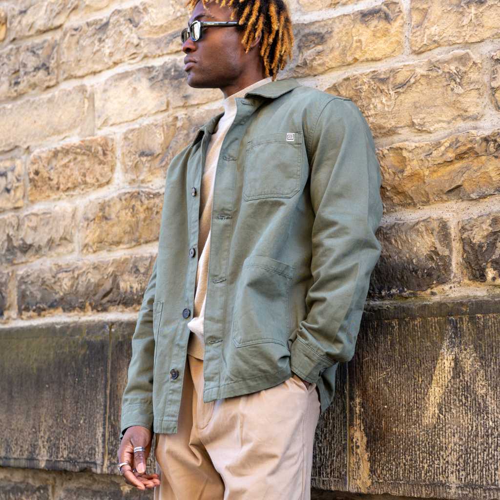 Man wearing a khaki paradaiza jacket and beige pants leaning against a stone wall