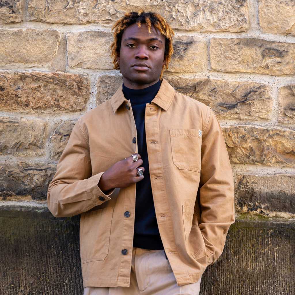 Man wearing a Tan paradaiza worker  jacket standing against a stone wall