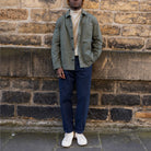 A person stands outdoors against a stone wall wearing an olive green utility-style jacket over a cream turtleneck, navy pants, and white sneakers.