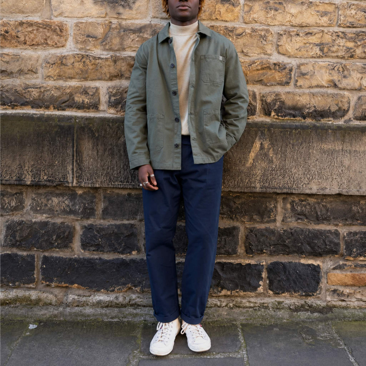 A person stands outdoors against a stone wall wearing an olive green utility-style jacket over a cream turtleneck, navy pants, and white sneakers.