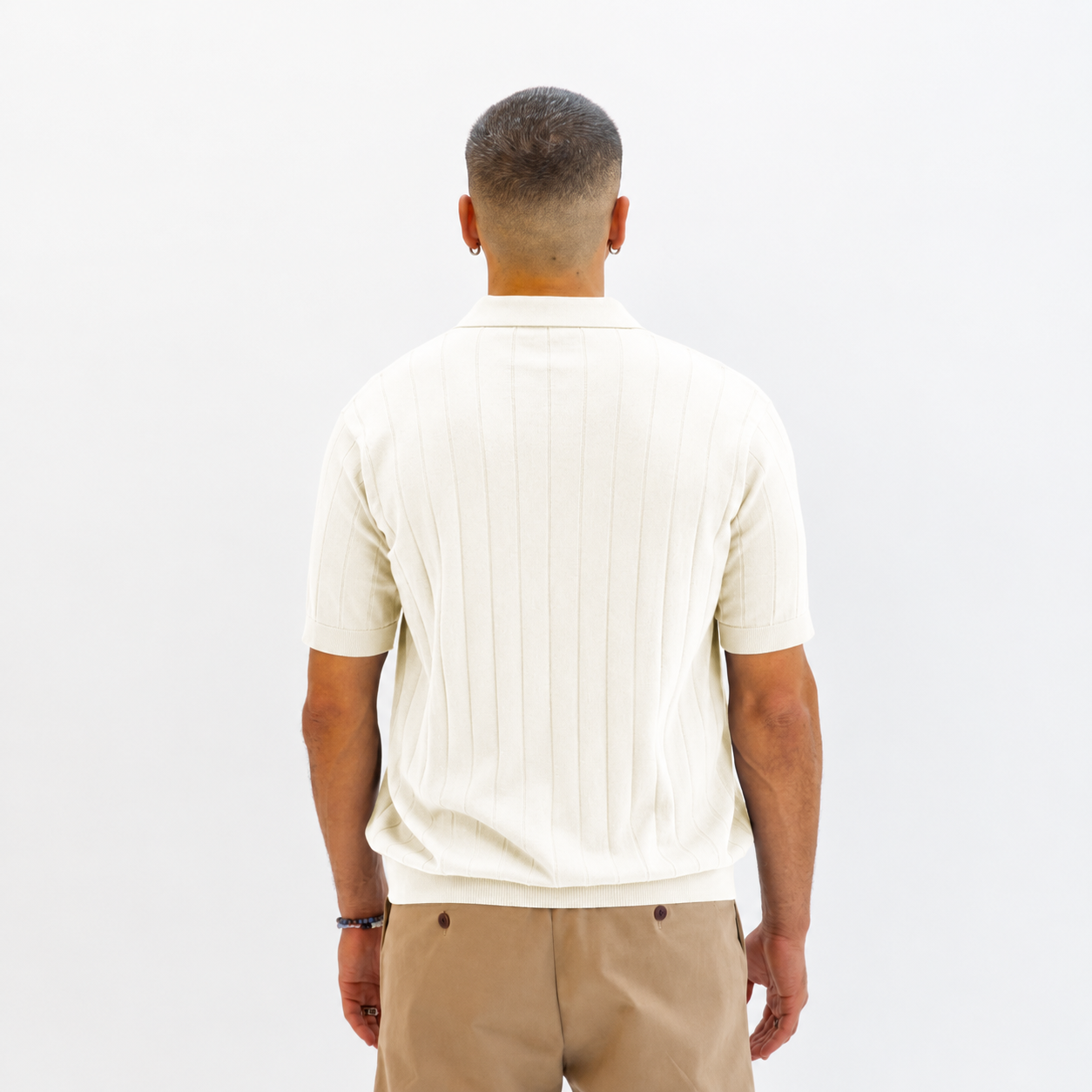 A man viewed from behind wearing a Paradaiza cream-colored short-sleeve polo shirt with vertical ribbed texture and tan trousers, set against a plain white studio background.