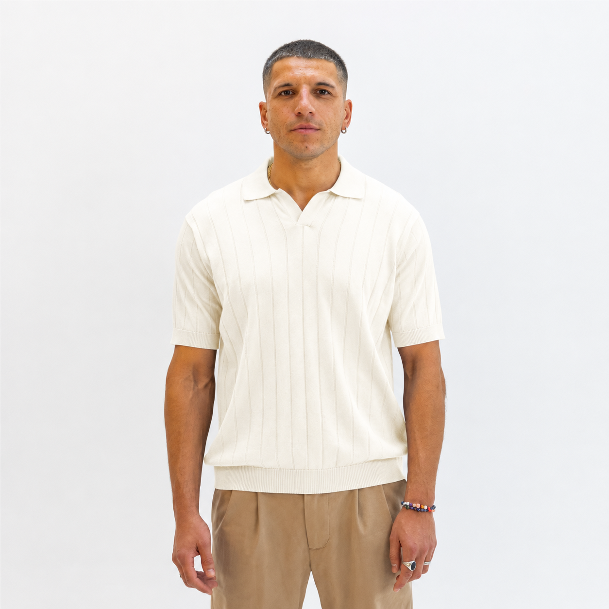 A man wearing a Paradaiza cream-colored short-sleeve knit polo shirt with vertical ribbed texture, paired with beige trousers, standing against a plain white studio background.