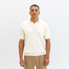A man wearing a Paradaiza cream-colored short-sleeve knit polo shirt with vertical ribbed texture, paired with beige trousers, standing against a plain white studio background.