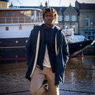 A man wearing a navy blue hooded jacket with cream fleece lining, a dark shirt, and beige trousers stands in a harbor in front of a large boat and brick buildings.