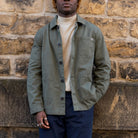 A man wearing an olive green utility overshirt over a cream turtleneck and navy trousers, standing against a rustic stone wall.