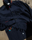 A dark navy blue jacket with a high collar, button front, and multiple pockets, laid flat on a textured woven surface. The jacket features small red embroidered details and a colorful patterned fabric accent visible inside a pocket.