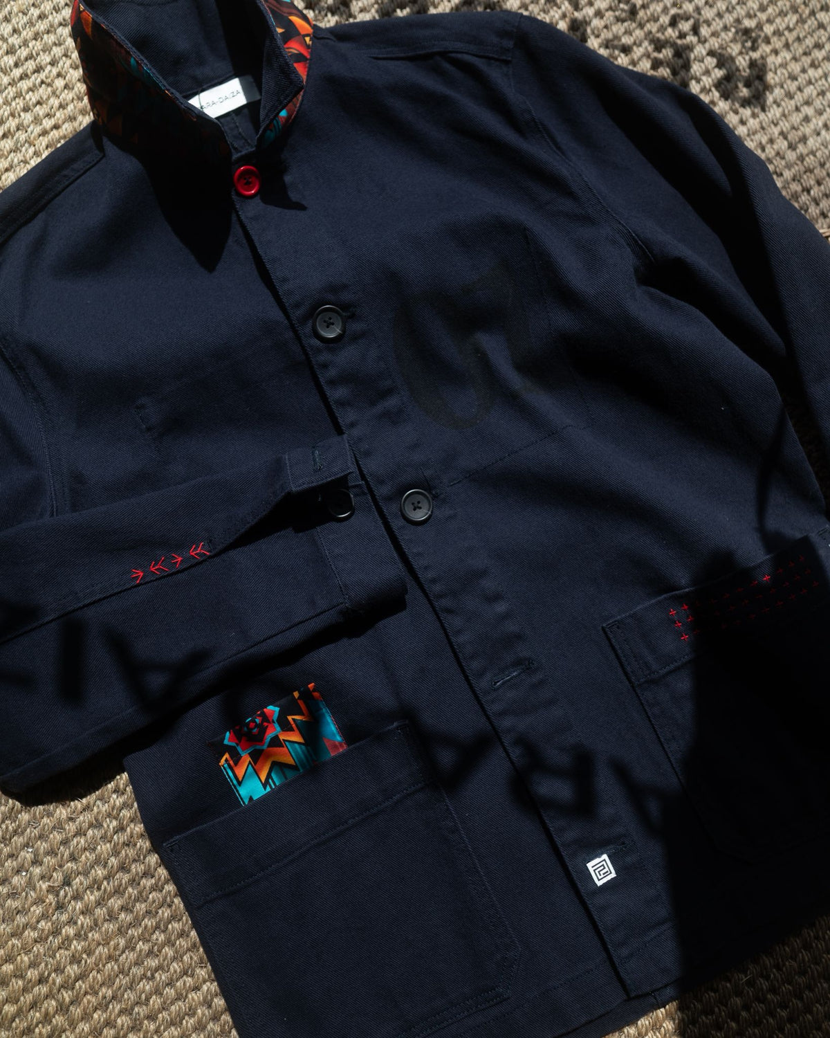 A dark navy blue jacket with a high collar, button front, and multiple pockets, laid flat on a textured woven surface. The jacket features small red embroidered details and a colorful patterned fabric accent visible inside a pocket.