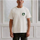 Man wearing a paradaiza  white t-shirt with a logo against a white paneled wall.
