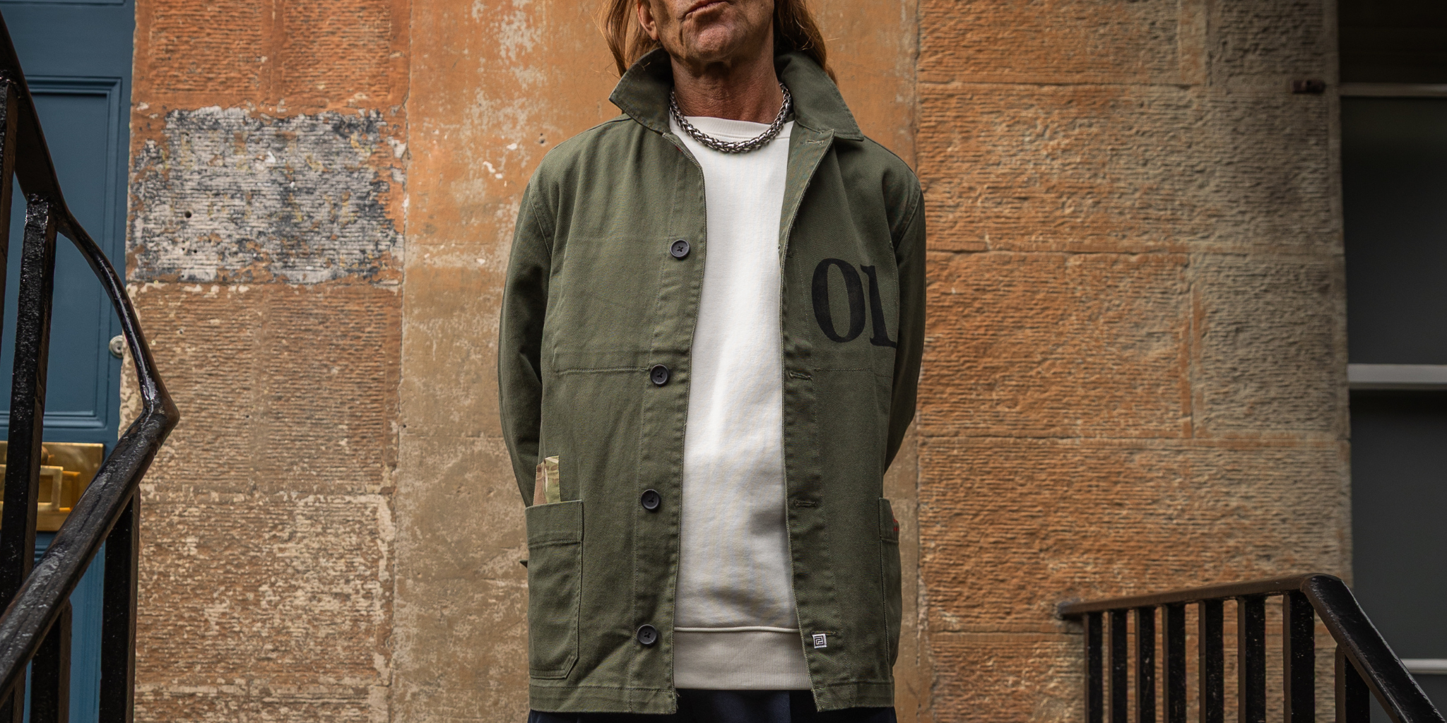 Person wearing a khaki PARA-DAIZA jacket with '01' branding against a stone wall in Edinburgh.