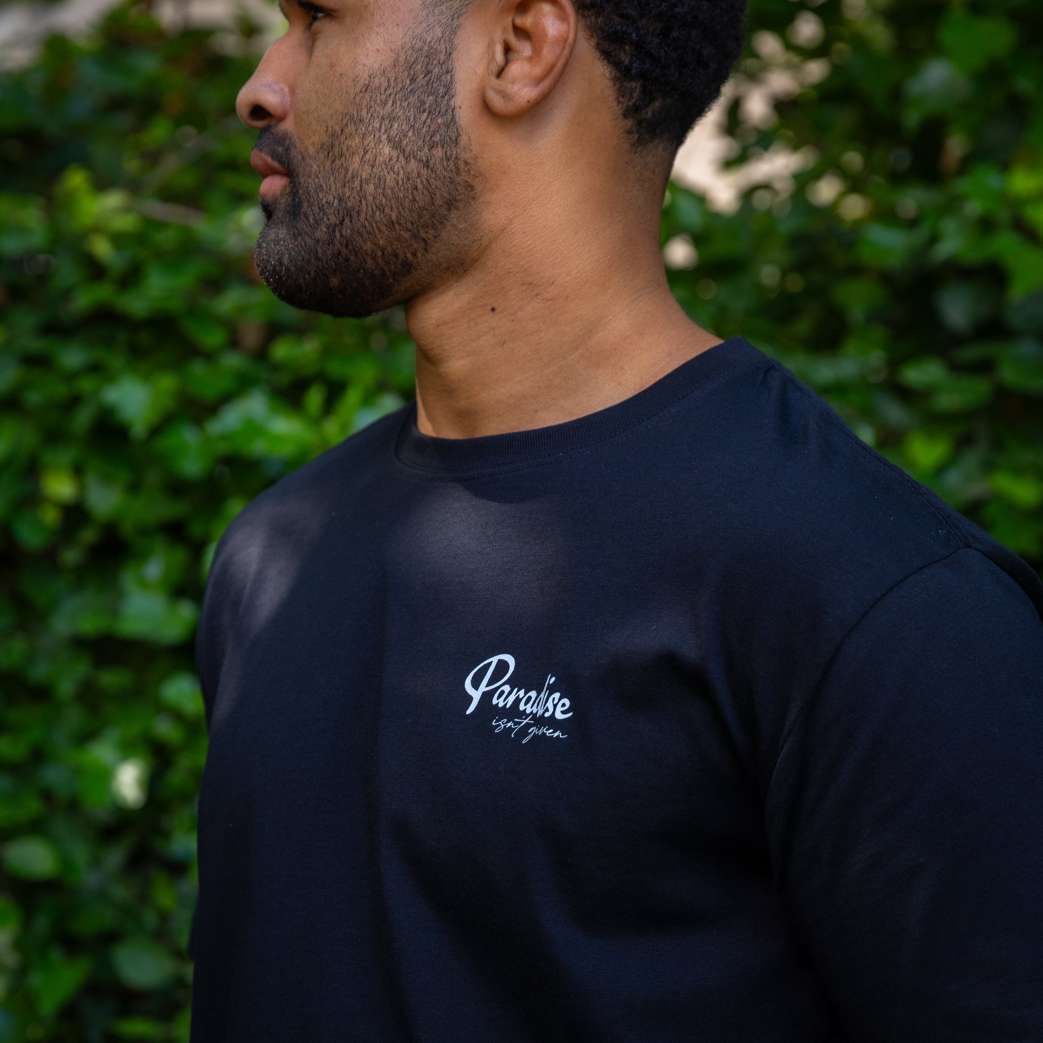 Man wearing a black paradaiza t-shirt with 'Paradise' logo against a green leafy background