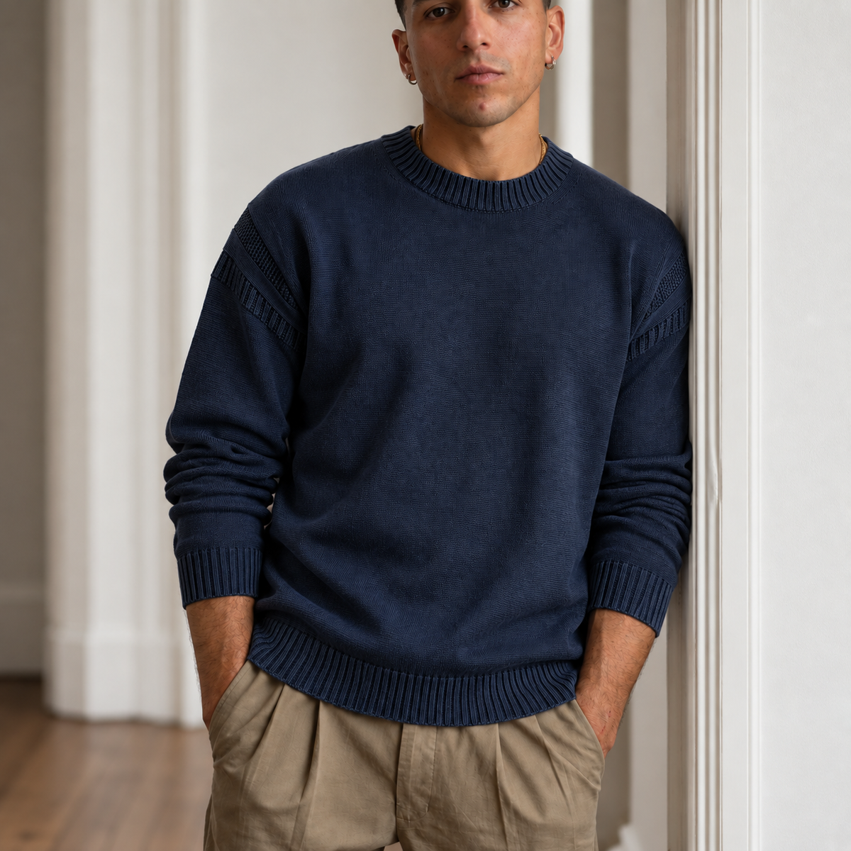 A man wearing a paradaiza navy blue crewneck sweater with textured knit details on the shoulders and cuffs, paired with khaki pants, standing in a doorway.